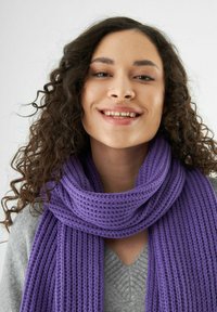 Jonge vrouw met krullend haar die glimlacht, gekleed in een dikke paarse gebreide sjaal en een lichtgrijze trui tegen een plain witte achtergrond.