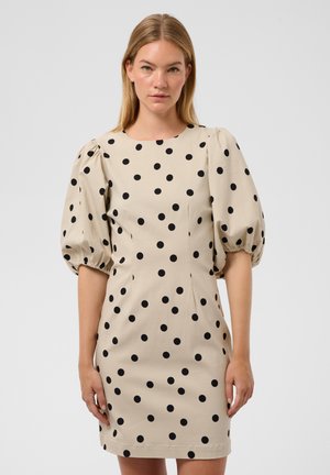Femme portant une robe beige à pois noirs avec des manches bouffantes, debout devant un fond uni gris clair.