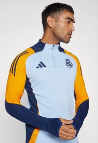 Világoskék, félig cipzáras pulóver narancssárga és sötétkék részletekkel. Rugalmas anyagból készült, az Adidas logójával és a Real Madrid emblémájával.