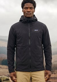 Mees seisab väljas musta kapuutsiga lukuga jope ja beežide pükstega, taustal pilvine ja künklik maastik.