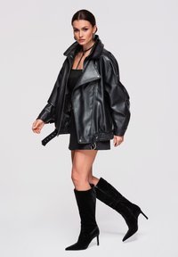 Vrouw draagt een oversized zwarte leren jas, een zwarte minirok en zwarte suède kniehoge laarzen met hakken, staande tegen een effen achtergrond.