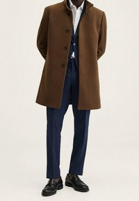 Uomo che indossa un cappotto marrone sopra un completo blu, una camicia bianca e scarpe eleganti nere, in piedi con le mani dietro la schiena su uno sfondo semplice.