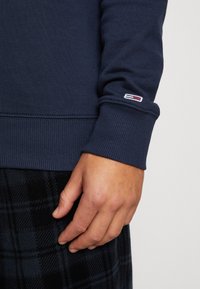 Sudadera azul marino con puños acanalados, que presenta un pequeño parche de marca. Mano descansando sobre una tela de cuadro oscuro abajo, destacando el contraste de texturas.