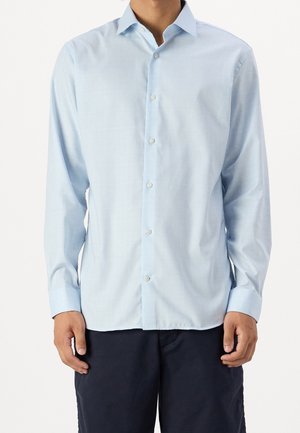 Mężczyzna ubrany w jasnoniebieską, zapinaną na guziki koszulę z długim rękawem i ciemne spodnie, stojący na jednolitym białym tle.