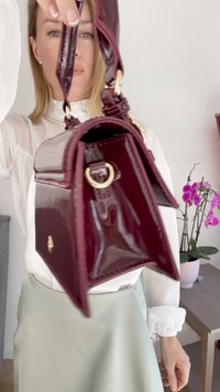 Magenta patentlæder håndtaske med gul hardware, rektangulær form, klapdesign og justerbar rem, holdt af en person.
