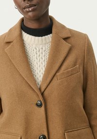 Blazer en mélange de laine marron avec des revers à cran, fermeture à un bouton et une poche. Porté par-dessus un pull crème épais avec une texture côtelée.