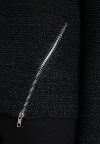 Svart stickad tröja med en ficka med dragkedja på sidan, i strukturerad tyg och med ribbad nederkant. Dragkedjan  är silverfärgad och synlig.