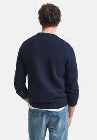 Mörkblå tröja med en texturerad, rutigt mönster, rundad halsringning och ribbade ärmslut, tillsammans med ljusblå jeans.