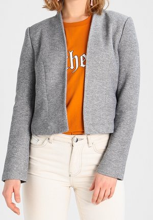 Frau trägt ein graues Blazer mit offenem Vorderteil über einem orangefarbenen T-Shirt mit Grafik und hoch taillierte hellbeige Hose.