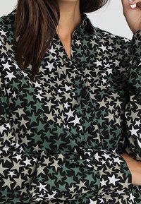 Camisa de manga larga con base negra y patrones de estrellas en verde, blanco y gris. Delantero abotonado con cuello y corte holgado.