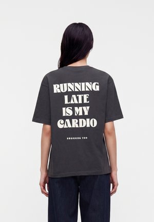 Personne aux longs cheveux foncés portant un t-shirt gris foncé avec le texte "RUNNING LATE IS MY CARDIO SHOPPING TOO" dans le dos.