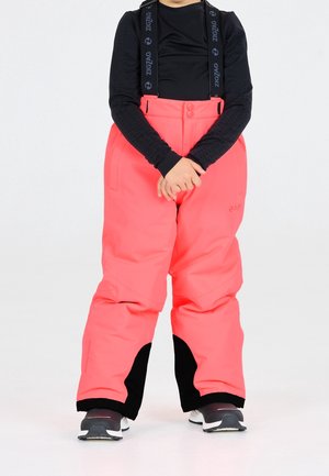 Dziecko ubrane w jaskrawo różowe spodnie narciarskie z czarnymi szelkami i czarną koszulkę z długim rękawem, stojące z złączonymi rękami na białym tle.