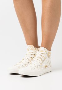 Hvide højhælede sneakers med blomstermønstrede gulddetaljer, lavet af canvasmateriale, rund tå og flade gummisåler, med traditionelt snørebåndsdesign.