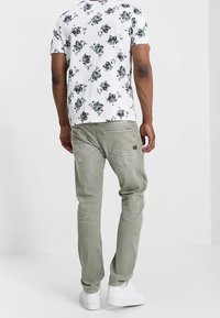 Tenue masculine comprenant un t-shirt blanc à motif floral et un pantalon vert olive, avec des poches visibles et des baskets blanches.
