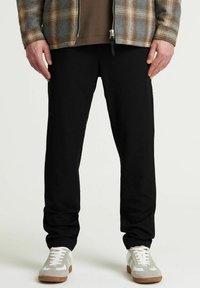Schwarze Jogginghosen aus weichem Stoff, mit lockerer Passform. Kombiniert mit einer karierten Jacke und grau-weißen Sneakers.