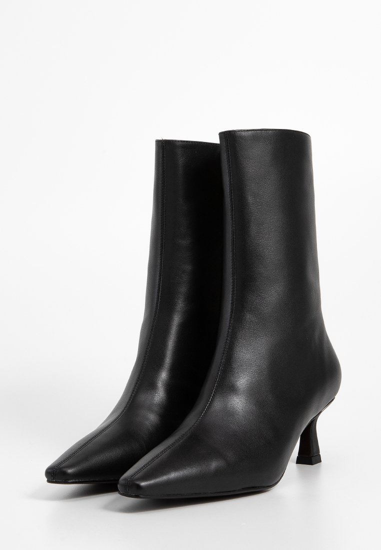 Paio di stivali in pelle nera fino al polpaccio con punta affusolata e tacchi kitten sottili su sfondo bianco.