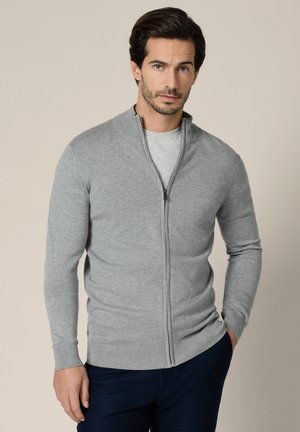 Mężczyzna w jasnoszarym swetrze na zamek, noszący szarą koszulę i ciemne spodnie, stojący na jednolitym beżowym tle z jedną ręką w kieszeni.