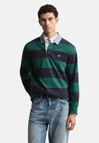 Zaļi un tumši zili svītrains polokrūzītes krekls ar garām piedurknēm un apkakli. Valkāts virs gaiši zilas pogām aiztaisāmas krekla. Ikdienas džinsa džinsi ar redzamu jostu.