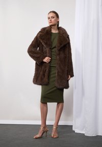 Manteau en fausse fourrure marron avec un large col, associé à une robe verte olive près du corps. La mannequin porte des talons dorés à lanières. Fond blanc et sol gris.
