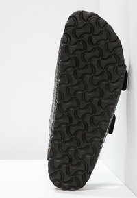 Semelle noire de sandale avec motif circulaire texturé, vue par le dessous, sur un fond blanc en angle.