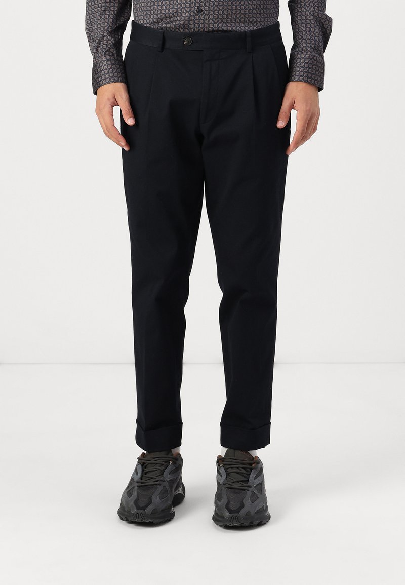 Black pleated trousers with a tailored fit, made of lightweight fabric. Cuffed hems, paired with textured gray sneakers featuring a mesh design.