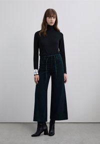 Pantalon large en velours côtelé vert foncé, associé à un haut noir ajusté à manches longues. Bracelet argenté au poignet, bottes noires à talons.