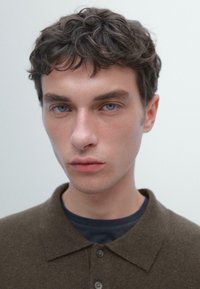 Giovane uomo con capelli corti ricci castani e occhi blu indossa una camicia scura con colletto, guardando direttamente in camera su uno sfondo semplice e chiaro.