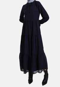 Marineblå, langærmet maxikjole med lagdelt design, samlet skørt og snore ved halsudskæringen, med gennemsigtig kantdetalje. Sorte ankelstøvler.