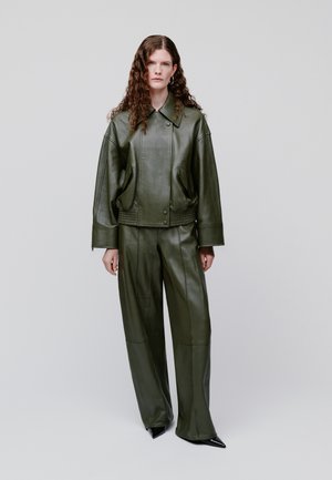 Γυναίκα με μακριά σγουρά μαλλιά που φοράει σκούρο πράσινο δερμάτινο μπουφάν και αντίστοιχο παντελόνι με φαρδιά μπατζάκια, στέκεται μπροστά σε έναν απλό φόντο.