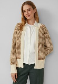 Strickjacke mit braunem und cremefarbenem Muster, gerippten Bündchen und offener Vorderseite. Tragen über einem weißen Hemd und grünen Hosen, strukturierter Stoff.