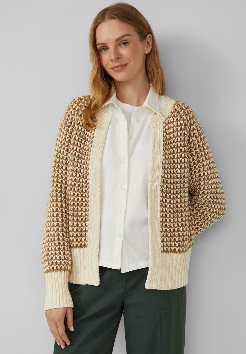 Strickjacke mit braunem und cremefarbenem Muster, gerippten Bündchen und offener Vorderseite. Tragen über einem weißen Hemd und grünen Hosen, strukturierter Stoff.