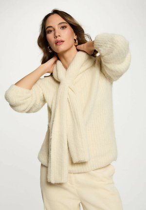 Maglione color crema con una morbida texture a coste, caratterizzato da un dettaglio con foulard annodato attorno al collo e ampie maniche arrotolate.