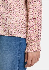Blusa a maniche lunghe con un motivo circolare in rosa, crema e marrone, con polsini arricciati e una consistenza del tessuto morbida. Abbinata a un paio di jeans.