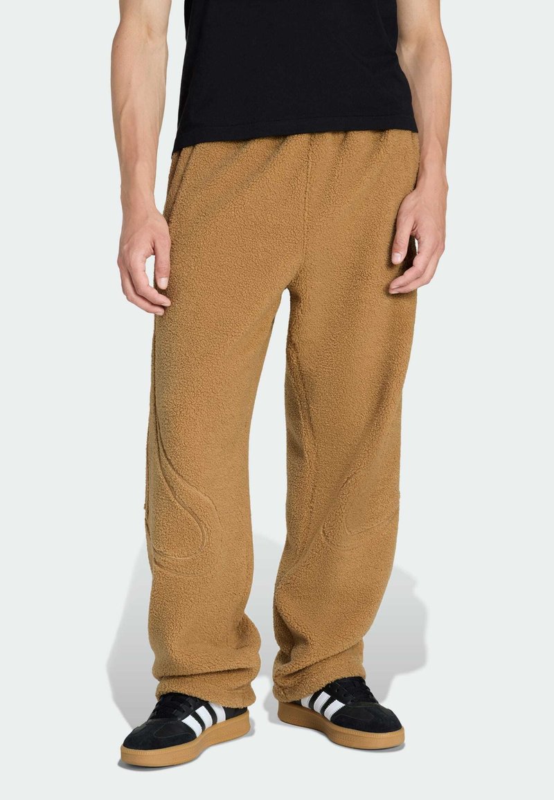 Hellbraune Fleece-Hosen mit strukturierter Oberfläche, lockerer Passform und dekorativen Nähten. Tragen mit schwarzen Schuhen mit weißen Streifen.