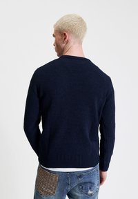 Granatowy sweter dziany z długimi rękawami, okrągłym dekoltem i ściągaczami. Nosi się go na podartych dżinsach z łatą w wzór houndstooth.