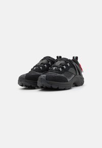 Black athletic shoes made of mesh and suede, featuring patterned detailing, robust rubber soles, and a distinctive red accent.