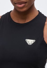 Κοντινό πλάνο ενός ατόμου που φοράει μαύρο αμάνικο τοπ με ένα λευκό στυλιζαρισμένο λογότυπο κεφαλιού πάνθηρα στο στήθος.