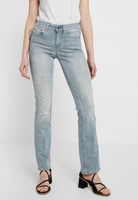 Persona vestida con unos jeans rectos de tiro alto en azul claro, una blusa sin mangas blanca, sandalias negras de tacón cuadrado con tiras, y con tatuajes visibles en los brazos.