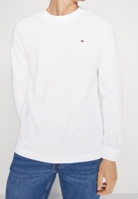 Vit långärmad sweatshirt i texturerad tyg. Har en rund halsringning och en liten broderad logotyp på bröstet. Bärs med blå jeans.