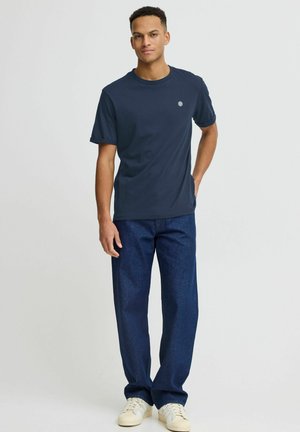 Marineblaue Baumwoll-T-Shirt mit kurzen Ärmeln, Rundhalskragen und kleinem Logo. Kombiniert mit geraden, dunklen Jeans und weißen Sneakers.