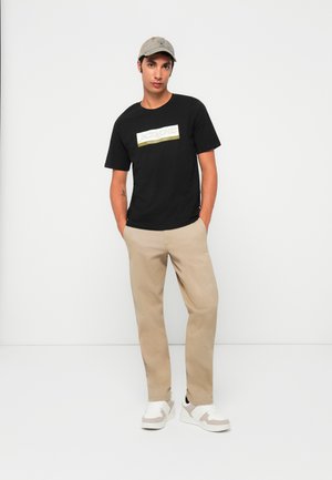 Young man wearing a beige cap, black Jack & Jones t-shirt, beige pants, and white sneakers, standing with hands in pockets against white background.