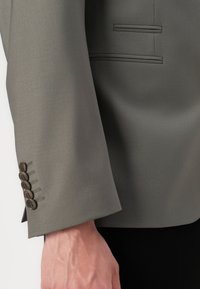 Detalle de la manga de un blazzer gris con textura suave, sutil brillo y cinco botones marrones en el puño, que presenta acentos de costura ocultos.