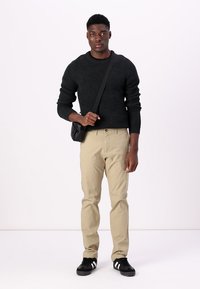 Pull noir texturé, pantalon beige clair et baskets noires avec des rayures blanches. Le mannequin tient un petit sac bandoulière noir.