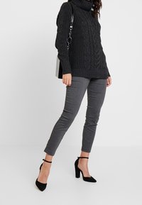 Zwarte gebreide coltrui met kabelpatroon, gecombineerd met grijze slim-fit pantalon en zwarte hoge hakken met enkelbandjes.