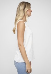 Wit mouwloos topje met een gestructureerd patroon, ronde halslijn en een licht asymmetrische zoom, gecombineerd met lichtblauwe spijkerbroek.