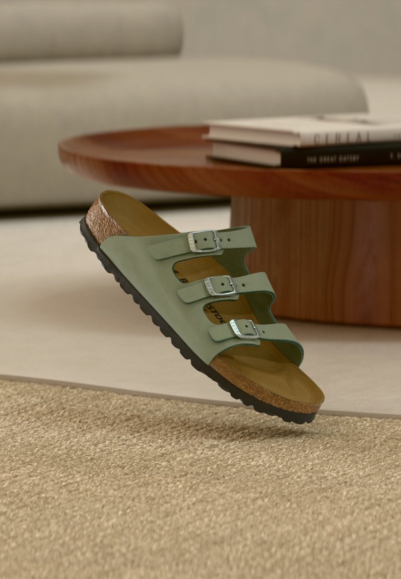 Πράσινο σανδάλι με τρεις ρυθμιζόμενους ιμάντες, ασημί αγκράφες, πάτο φτιαγμένο από φελλό και υφασμένη μαύρη ελαστική σόλα, τοποθετημένο πάνω σε υφαντό υπόστρωμα.