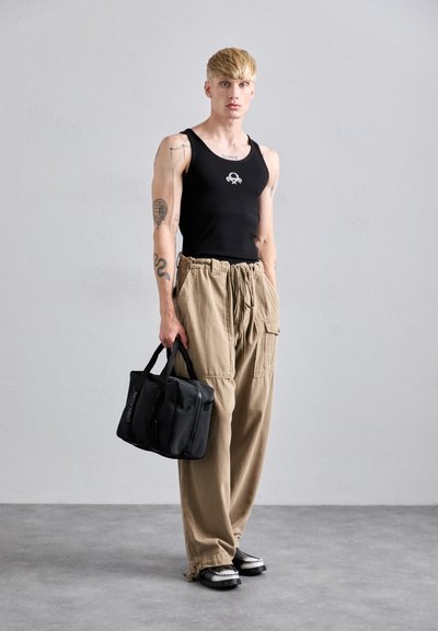 Tânăr cu tatuaje poartă un top fără mâneci negru, pantaloni cargo bej, pantofi negri și ține o geantă neagră Moschino lângă un zid gri simplu.