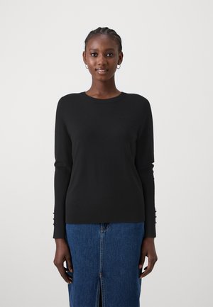 Black knitted sweater with a round neckline, long sleeves featuring gold button details on the cuffs. Worn over a denim skirt.
