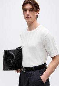 Νεαρός άνδρας που φοράει λευκό κοντομάνικο πουκάμισο και σκούρο παντελόνι κρατά μια μαύρη υφαντή δερμάτινη τσάντα στο ύψος της μέσης.