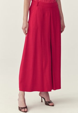 Woman wearing a long, bright red skirt with a front slit, paired with brown open-toe high-heeled mules, standing on a light background.